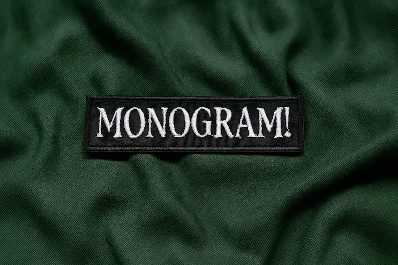 Pu&ograve; includere: Una toppa rettangolare nera con la scritta "MONOGRAM!" ricamata in bianco, su uno sfondo di tessuto verde scuro. La toppa &egrave; centrata e il testo &egrave; in grassetto, in un carattere stilizzato.