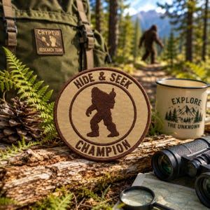 Pode incluir: Um emblema bordado circular apresentando uma silhueta castanha de uma criatura no centro com o texto HIDE & SEEK CHAMPION em letras castanhas sobre um fundo bege, rodeado por uma borda castanha escura, exibido num tronco de madeira.