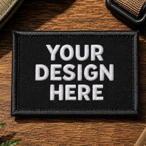 May include: Black rectangular patch with white embroidered text that reads "YOUR DESIGN HERE." The patch has a black border and is set against a wooden background. Other items in the image include a green plant and a brown strap.