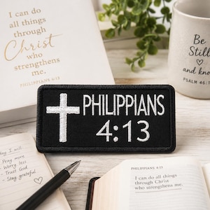May include: A black patch with white embroidered text that reads "PHILIPPIANS 4:13" and a white cross. A notebook, pen, and open book are also visible. A white mug with the text "Be Still and know" is in the background.