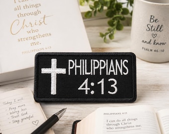 以下が含まれることがあります： 「PHILIPPIANS 4:13」と白い十字架が刺繍された白い文字の黒いパッチ。ノート、ペン、開いた本も見えます。「Be Still and know」というテキストが入った白いマグカップが背景にあります。