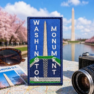 May include: A blue embroidered patch with the words "WASHINGTON MONUMENT" and an image of the Washington Monument. The patch is on a map with a camera and sunglasses in the background. Cherry blossoms and the Washington Monument are visible.
