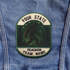 May include: Embroidered patch with a green and white border, featuring a silhouette of Bigfoot in a forest scene. The patch has the text "YOUR STATE" at the top and "TRACKER TEAM NAME" at the bottom, set against a denim background.