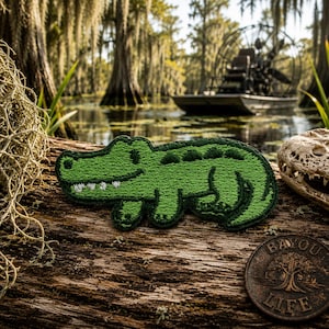 May include: A green embroidered alligator patch with white teeth, lying on a weathered wooden surface. The patch is a cartoon-style design. A bronze coin with the words "Bayou Life" is also visible. The background shows a swamp scene with an airboat.