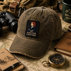 May include: A distressed olive green baseball cap featuring a rectangular patch with an illustration of a person with red hair and the text "QUESTION EVERYTHING" in white capital letters centered on the front panel of the hat.