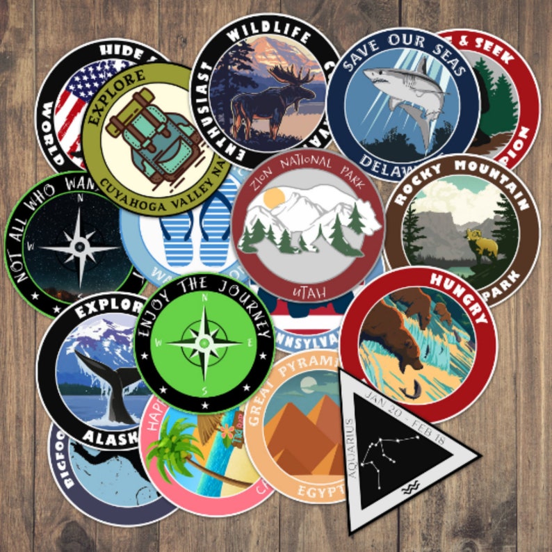 May include: A collection of 14 colorful round stickers with various designs, including a compass, a whale tail, a mountain range, a moose, a shark, a palm tree, and a compass. The stickers are all on a wooden background.