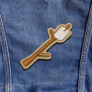May include: Embroidered patch featuring a marshmallow on a stick, in shades of white, brown, and gold. The patch is sewn onto a blue denim jacket. The design depicts a toasted marshmallow, a common camping treat.