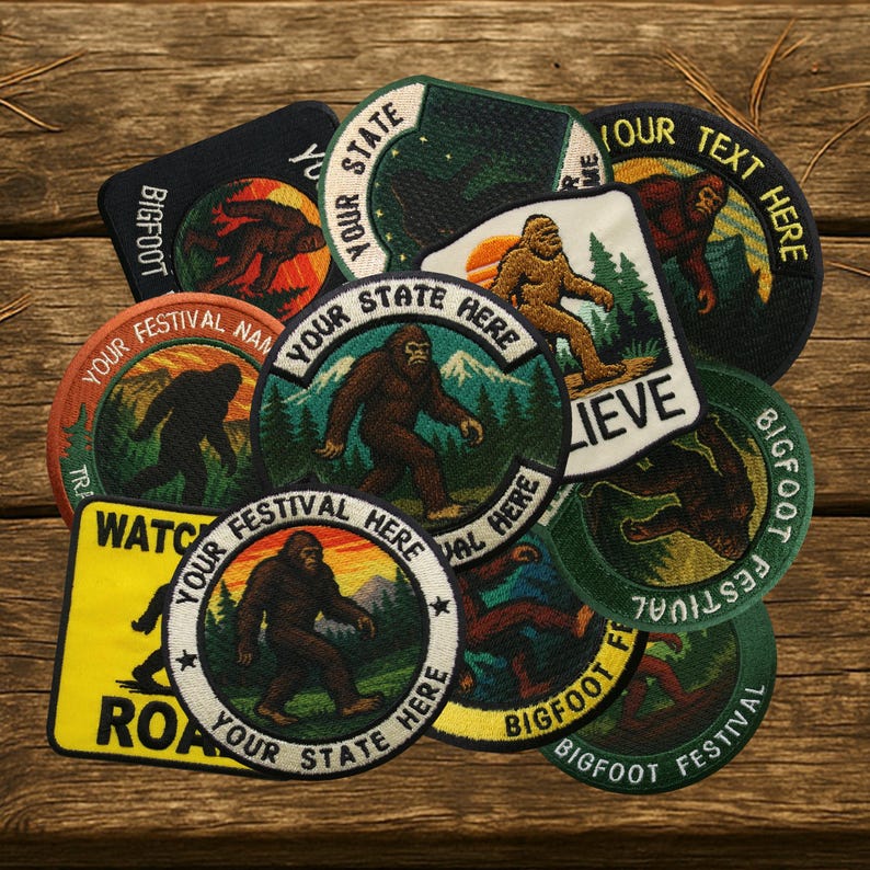 May include: A collection of Bigfoot-themed patches in various shapes and sizes. The patches feature images of Bigfoot, mountains, and text like "Your State Here" and "Bigfoot Festival." Colors include green, brown, orange, and black. The patches are arranged on a wooden surface.