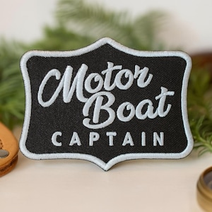 Könnte beinhalten: Schwarzer Aufnäher mit weiß gesticktem Text „Motor Boat Captain“. Der Aufnäher hat eine dekorative Bordüre. Ein Lederartikel, ein Kompass und Grünzeug befinden sich im Hintergrund.