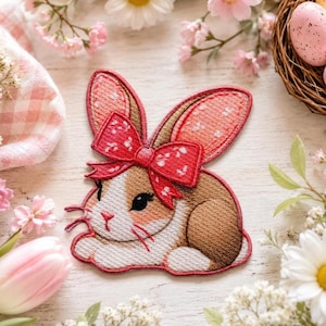 May include: An embroidered patch featuring a brown and white rabbit with long ears, wearing a large red bow with white polka dots. The rabbit is depicted in a resting position with its head turned to the side, showcasing detailed stitching.