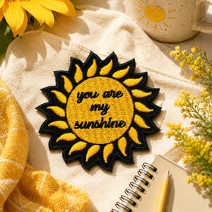 May include: A circular embroidered patch featuring a yellow sun design with black rays and the black text you are my sunshine centered in the middle. The patch has a thick black border and is crafted with detailed stitching throughout the design.