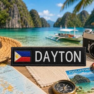 以下が含まれることがあります： フィリピンの国旗と白で刺繍された「DAYTON」という名前が入った黒いパッチ。麦わら帽子、ヴィンテージカメラ、地図、コンパス、ポストカードとともに表示され、旅行をテーマにしています。