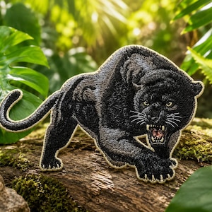 May include: An embroidered patch featuring a black panther in a prowling, aggressive stance with its mouth open, showing teeth. The panther has detailed black thread work, yellow eyes, and a light-coloured border outlining the entire animal shape.