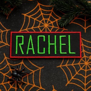 May include: A black rectangular patch with the name "RACHEL" embroidered in green letters, outlined in red. The patch is set against a black background with orange spiderwebs and a small black spider. Green pine branches are in the background.