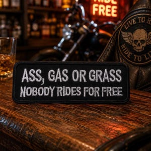 May include: A rectangular black embroidered patch featuring white stitched text that reads, "ASS, GAS OR GRASS NOBODY RIDES FOR FREE." The patch has a clean, finished border and is displayed on a dark wooden surface with a blurred motorcycle background.