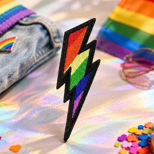 May include: An embroidered lightning bolt patch featuring a vibrant rainbow gradient of red, orange, yellow, green, blue, and purple threads. The design is outlined with a thick black border, creating a sharp contrast against the colorful interior.