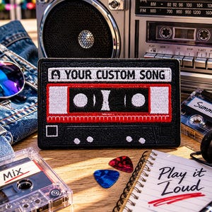 Könnte beinhalten: Ein schwarzer Aufnäher mit einem Kassettenband-Design und dem Text "A YOUR CUSTOM SONG". Das Bild zeigt einen Vintage-Boombox, Sonnenbrillen, Kassetten und einen Notizblock mit der Aufschrift "Play it Loud".