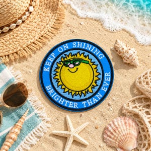 Puede incluir: Un parche redondo azul con el texto "KEEP ON SHINING BRIGHTER THAN EVER" que rodea un sol amarillo sonriente con gafas de sol verdes. El parche está en una playa de arena con un sombrero de paja, gafas de sol y conchas marinas.