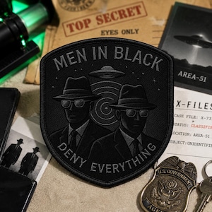 May include: Black embroidered patch with the text "MEN IN BLACK DENY EVERYTHING" and an illustration of two figures in suits and hats. The patch also features a UFO design. Other items in the image include a badge, keys, and documents.