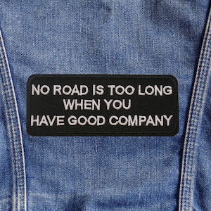 May include: A black rectangular patch with white embroidered text that reads "NO ROAD IS TOO LONG WHEN YOU HAVE GOOD COMPANY" is sewn onto a blue denim garment. The patch is centered and the denim has visible seams.