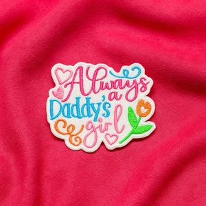 May include: Embroidered patch with the text "Always a Daddy's Girl" in pink, blue, and orange, with heart and flower accents. The patch is on a bright pink fabric background.