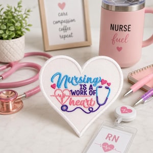 May include: A heart-shaped embroidered patch with the text "Nursing is a work of heart" in blue and pink. A pink travel mug with "NURSE fuel" printed on it. A stethoscope and a badge holder are also visible.