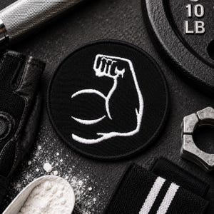 May include: A circular black embroidered patch featuring a white stitched outline of a flexed bicep arm. The patch is surrounded by gym equipment including a weight plate marked 10 LB, a metal barbell, a black glove, and a white powder scoop.