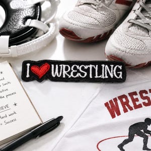 May include: A still-life image featuring wrestling gear and motivational items. Includes a white wrestling shirt with a red graphic, a black and white patch that says "I ❤️ WRESTLING", a notepad, pen, wrestling shoes, and a head guard.