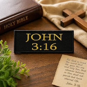May include: A black rectangular patch with the text "JOHN 3:16" embroidered in gold. The image also includes a closed brown Bible, a wooden cross, and a handwritten note with text from John 3:16.