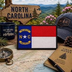 May include: A rectangular embroidered patch featuring the North Carolina state flag with a blue vertical stripe on the left containing a white star, the letters N and C, and two yellow ribbons with the dates May 20th 1775 and April 12th 1776.