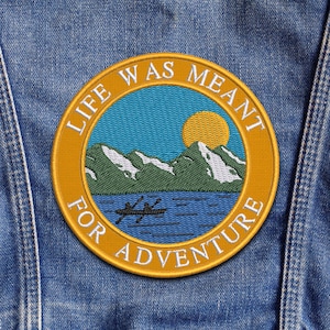 Könnte beinhalten: Runder, gestickter Aufnäher mit einer Berg- und Seeszene und dem Text "LIFE WAS MEANT FOR ADVENTURE". Der Aufnäher hat einen gelben Rand, blauen Himmel, grüne Berge und eine gelbe Sonne. Er ist auf eine Jeansjacke genäht.