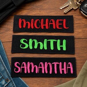 May include: Three black rectangular patches with embroidered names. The top patch reads "MICHAEL" in red script. The middle patch says "SMITH" in green script. The bottom patch says "SAMANTHA" in pink script.