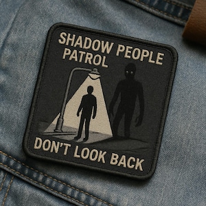 May include: Black square patch with the words "SHADOW PEOPLE PATROL DON'T LOOK BACK" in white. The patch features a silhouetted figure under a streetlight, with a shadow figure nearby. The patch is on a denim jacket.