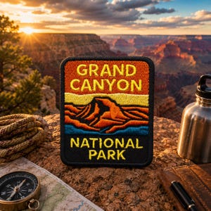 May include: A rectangular embroidered patch featuring the text GRAND CANYON NATIONAL PARK in yellow letters. The design depicts a stylized landscape with orange, yellow, and blue layers representing canyon rock formations against a black border.