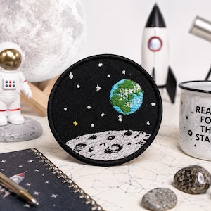 May include: A round black patch with an embroidered design of the Earth, moon, and stars. The patch is surrounded by space-themed items, including a rocket, astronaut figurine, and a mug that says "Reach for the Stars."