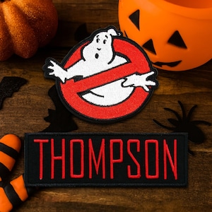 May include: A Halloween-themed patch set featuring a Ghostbusters logo patch and a custom name patch that reads "THOMPSON" in red letters on a black background. Decorative pumpkins and spider accents complete the spooky scene.