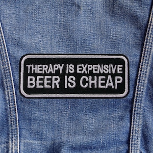 Puede incluir: Un parche rectangular negro con texto blanco que dice "THERAPY IS EXPENSIVE BEER IS CHEAP". El parche está bordeado con costuras blancas y está adherido a una prenda de mezclilla azul.