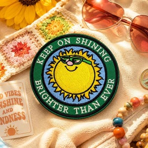 May include: A circular embroidered patch featuring a yellow sun with a smiling face and green sunglasses. The sun is set against a light blue background, surrounded by a dark green border with the white text KEEP ON SHINING BRIGHTER THAN EVER.