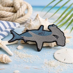 Op de afbeelding: Een geborduurde patch in de vorm van een hamerhaai met een blauw en grijs kleurenschema en een zwarte gestikte rand. Het haai-ontwerp bevat een zichtbare mond met kleine witte tanden en een kenmerkende brede kop, gemaakt met gedetailleerd stikwerk.