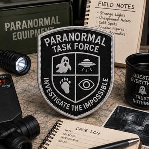 May include: A shield-shaped embroidered patch with a black background and silver stitching. It features the text PARANORMAL TASK FORCE and INVESTIGATE THE IMPOSSIBLE, with icons of a ghost, a UFO, a paw print, and an eye centered in the shield.