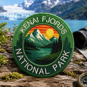 May include: A circular embroidered patch featuring a landscape of snow-capped mountains, a forest, and a lake under a yellow sun. The border is dark green with the white text KENAI FJORDS NATIONAL PARK stitched in a circular pattern around the center.
