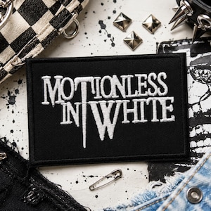 Puede incluir: Un parche rectangular negro bordado con el texto blanco Motionless In White en una fuente estilizada. El parche tiene un borde cosido limpio y está centrado, mostrando letras en negrita con una línea vertical que sale de la letra T.