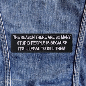 May include: A black rectangular patch with white embroidered text: "THE REASON THERE ARE SO MANY STUPID PEOPLE IS BECAUSE IT'S ILLEGAL TO KILL THEM." The patch is attached to a blue denim material, typical of jeans.