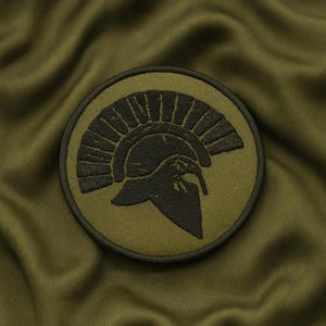 Könnte beinhalten: Runder, olivgrüner Aufnäher mit einem dunkelgrünen, gestickten Spartanerhelm-Design. Der Aufnäher zeigt eine detaillierte Darstellung des Helms, vor einem passenden olivgrünen Hintergrund. Geeignet für Kleidung oder Accessoires.
