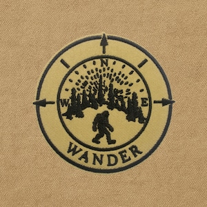 Peut inclure: Un écusson circulaire avec un design de boussole et le mot "WANDER". L'écusson présente une silhouette noire d'une créature ressemblant à Bigfoot, des arbres et des flèches directionnelles. Le fond est en tissu beige.