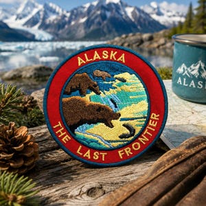 May include: A circular embroidered patch featuring a brown bear catching a fish in a blue river, surrounded by a red border with the yellow text ALASKA THE LAST FRONTIER. The design includes yellow and green landscape details within the circle.