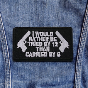 Peut inclure: Écusson rectangulaire noir avec texte brodé blanc et deux silhouettes de pistolets. Le texte se lit comme suit : « I WOULD RATHER BE TRIED BY 12 THAN CARRIED BY 6 ». L'écusson est présenté sur un fond en denim.