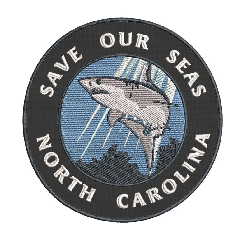 May include: Embroidered patch with a shark leaping out of the water. The patch is round with the text "SAVE OUR SEAS NORTH CAROLINA" in white on a black background. The shark is white and gray.