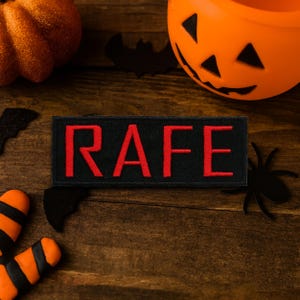Peut inclure: Un écusson noir avec le nom "RAFE" brodé en rouge. L'écusson est rectangulaire et entouré de décorations d'Halloween, dont une citrouille, des chauves-souris et une araignée, le tout sur une surface en bois.