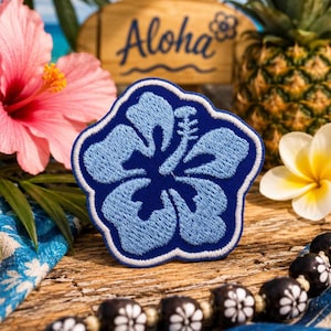 May include: A fabric patch featuring an embroidered light blue hibiscus flower on a dark blue background with a white border. The flower has five petals and a prominent stamen. The patch is shaped like the flower itself with a scalloped edge.